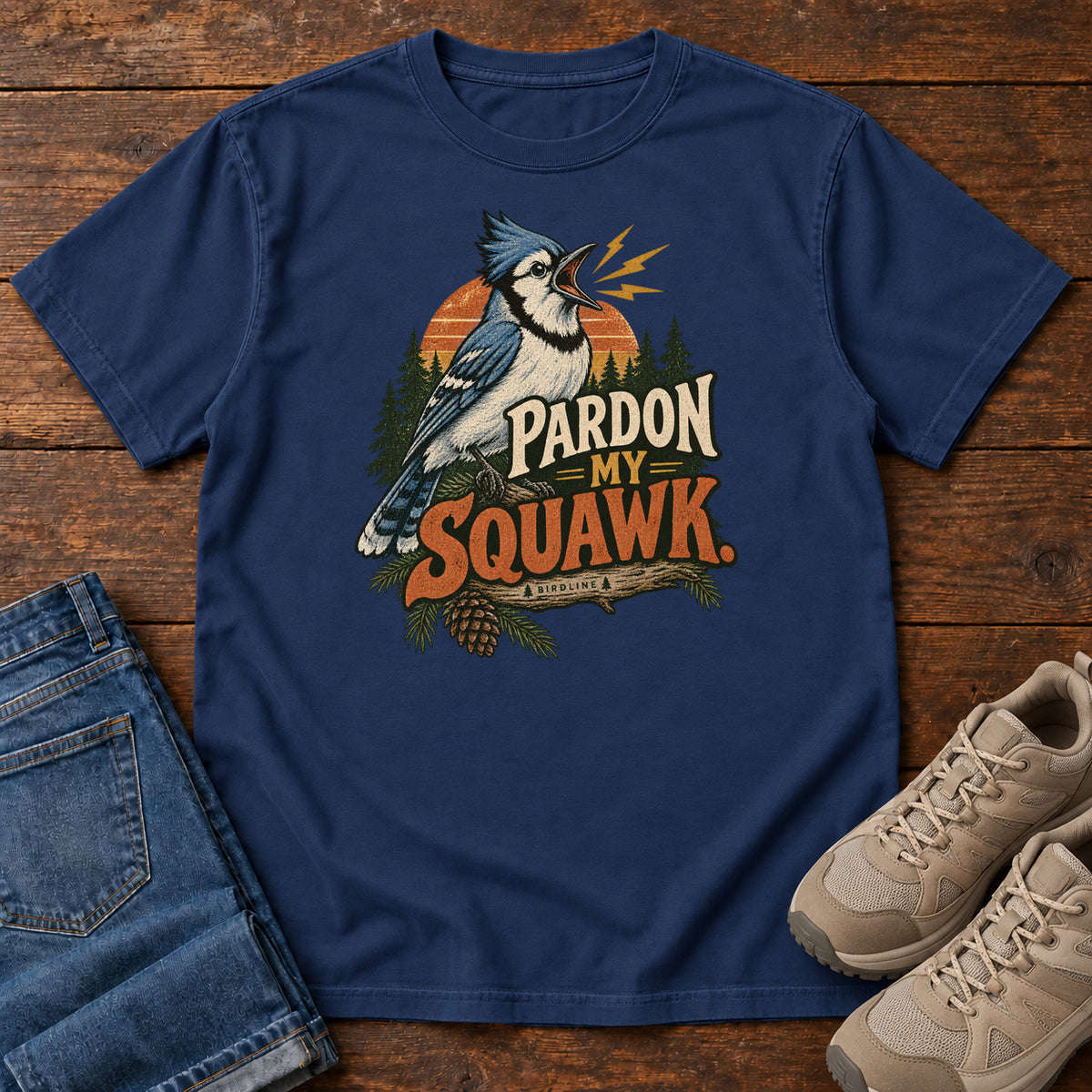 Pardon My Squawk T-Shirt in True Navy, front laid flat on wood, featuring a blue-and-white crested bird with an open beak, orange sound marks, pine trees and bold lettering.