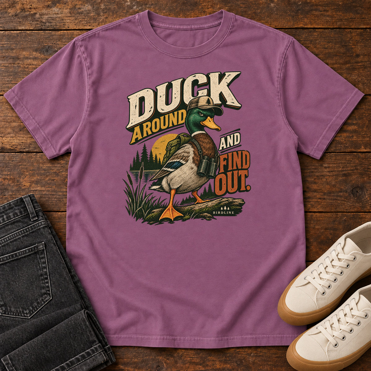 Berry Duck Around and Find Out T-Shirt with a green-headed duck in a hat and vest beside marsh reeds and bold lettering, front flat lay on wood.