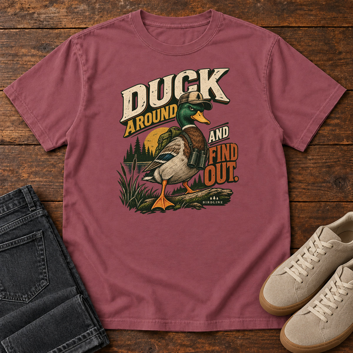 Brick Duck Around and Find Out T-Shirt with a green-headed duck in a hat and vest beside marsh reeds and bold lettering, front flat lay on wood.