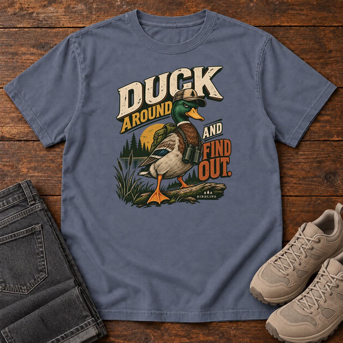 Denim Duck Around and Find Out T-Shirt with a green-headed duck in a hat and vest beside marsh reeds and bold lettering, front flat lay on wood.