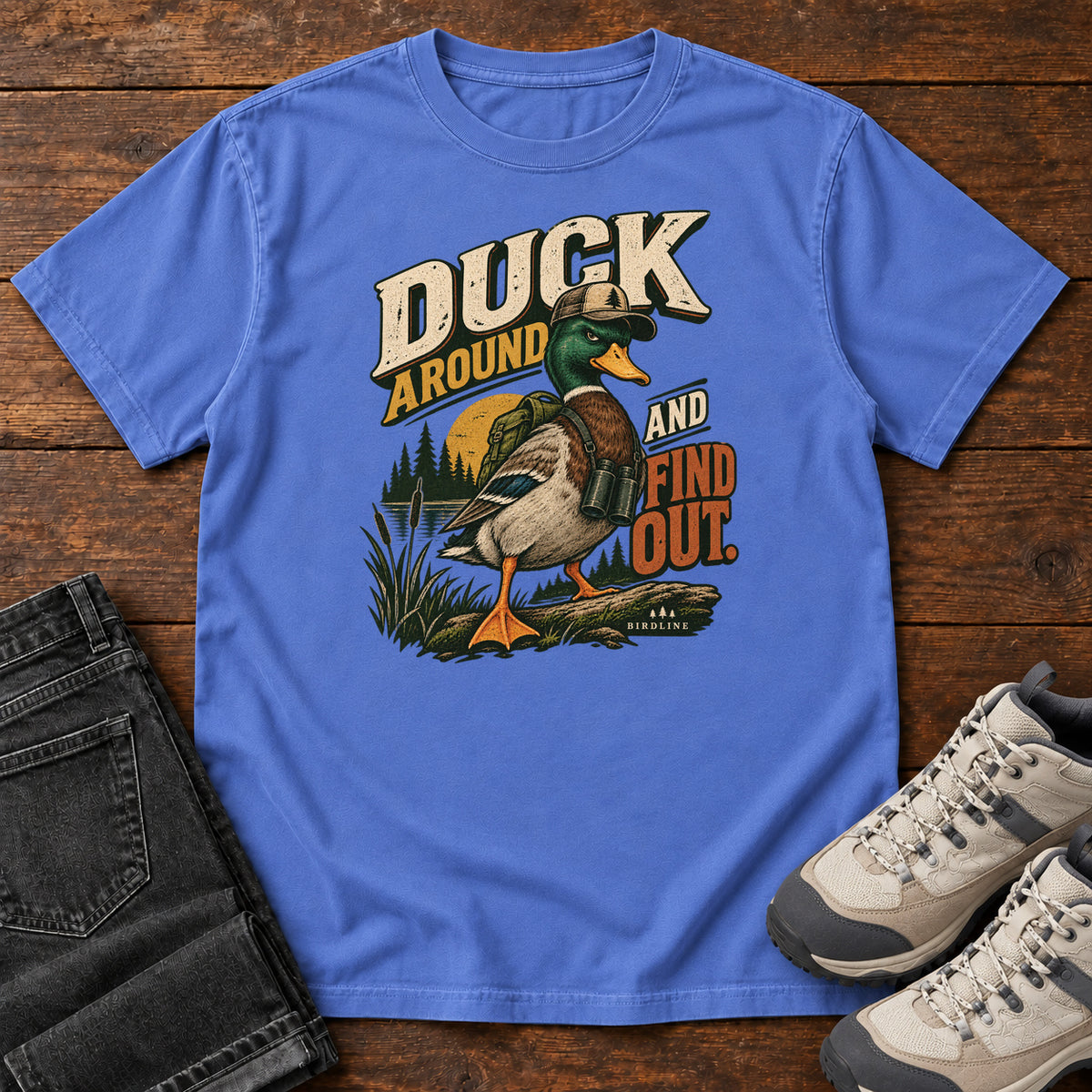 Mystic Blue Duck Around and Find Out T-Shirt with a green-headed duck in a hat and vest beside marsh reeds and bold lettering, front flat lay on wood.