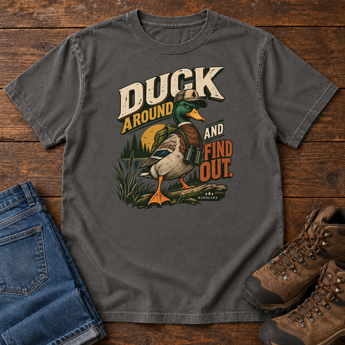 Pepper Duck Around and Find Out T-Shirt with a green-headed duck in a hat and vest beside marsh reeds and bold lettering, front flat lay on wood.