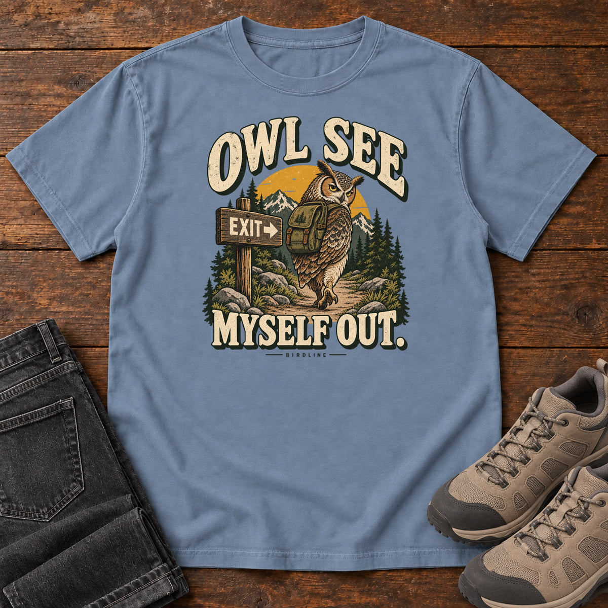 Blue Jean Owl See Myself Out T-Shirt with an owl wearing a backpack beside an Exit sign on a woodland trail; front flat lay on wood.