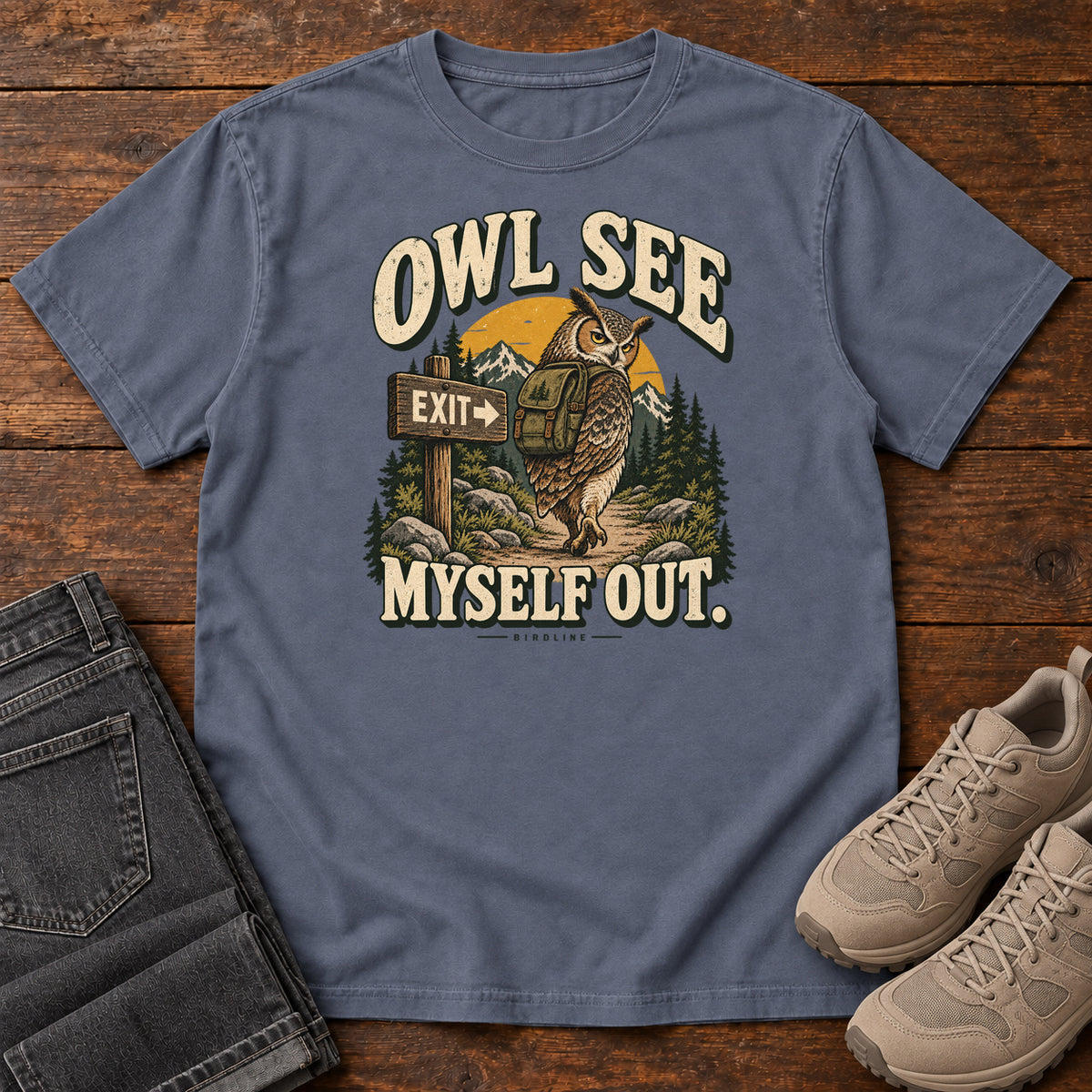 Denim Owl See Myself Out T-Shirt with an owl wearing a backpack beside an Exit sign on a woodland trail; front flat lay on wood.