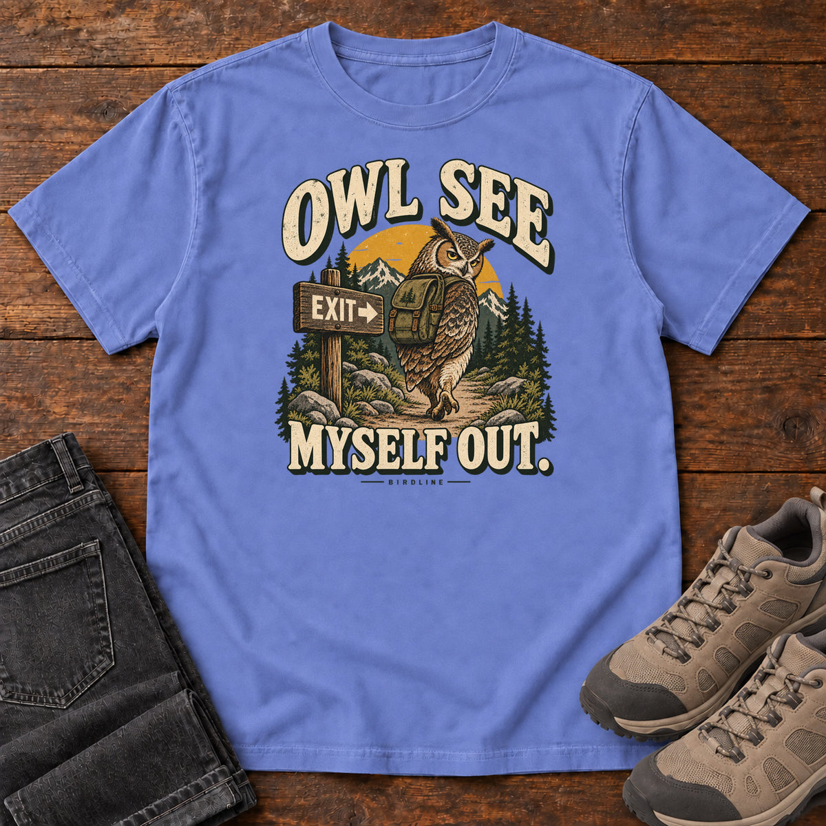 Flo Blue Owl See Myself Out T-Shirt with an owl wearing a backpack beside an Exit sign on a woodland trail; front flat lay on wood.