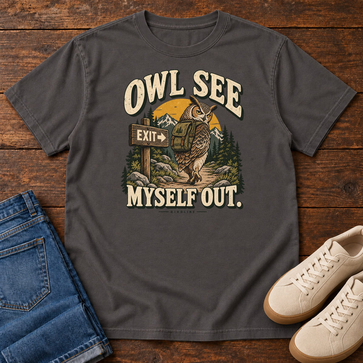 Graphite Owl See Myself Out T-Shirt with an owl wearing a backpack beside an Exit sign on a woodland trail; front flat lay on wood.
