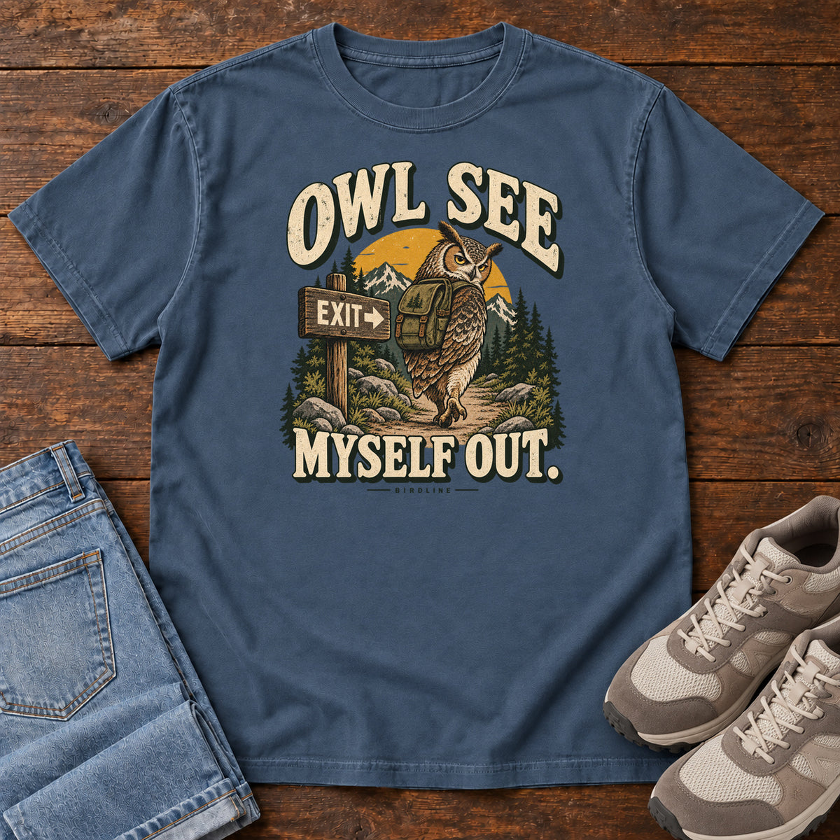 Midnight Owl See Myself Out T-Shirt with an owl wearing a backpack beside an Exit sign on a woodland trail; front flat lay on wood.