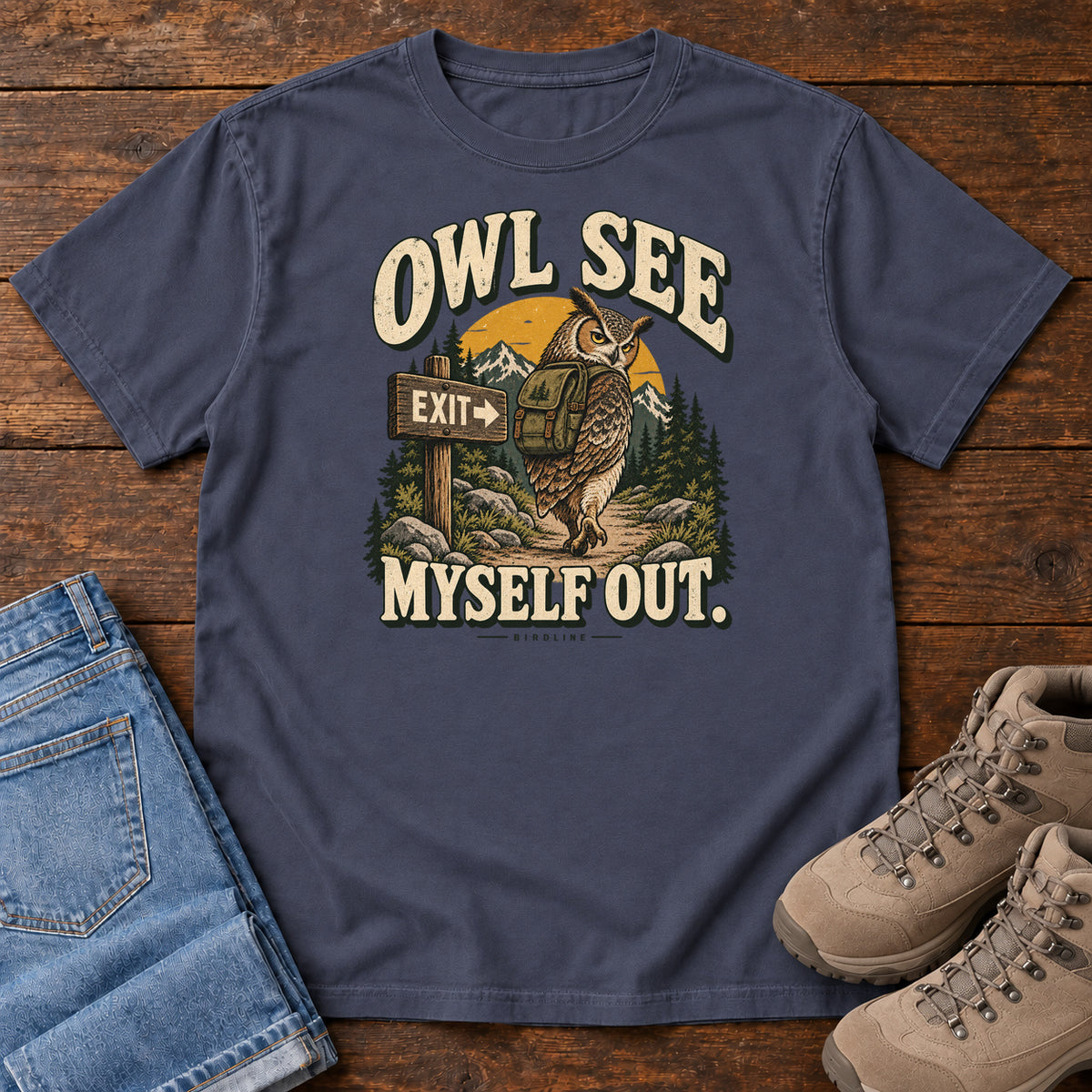 Navy Owl See Myself Out T-Shirt with an owl wearing a backpack beside an Exit sign on a woodland trail; front flat lay on wood.