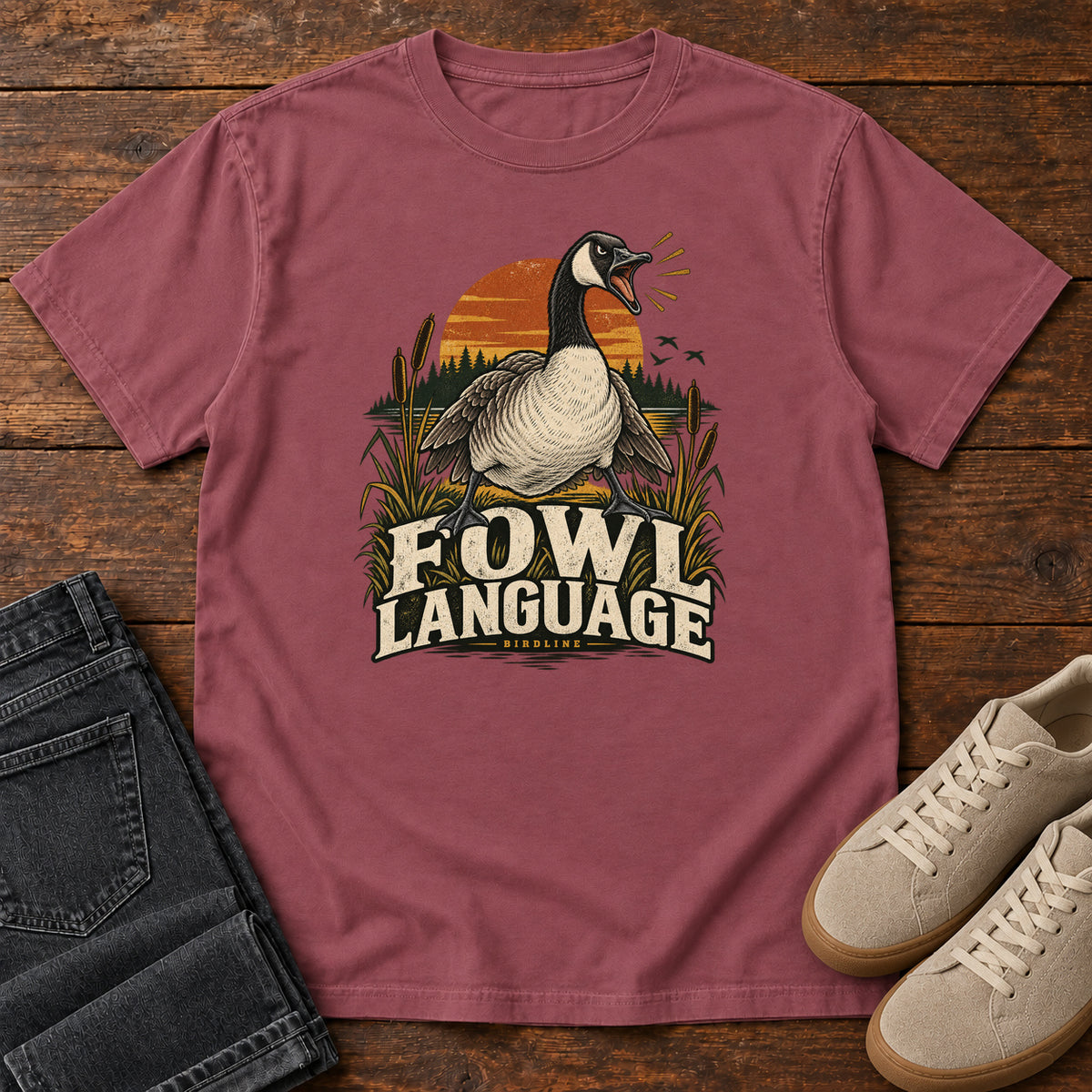 Brick Fowl Language T-Shirt with an open-beaked goose among reeds against an orange sunset; front flat lay on wood.