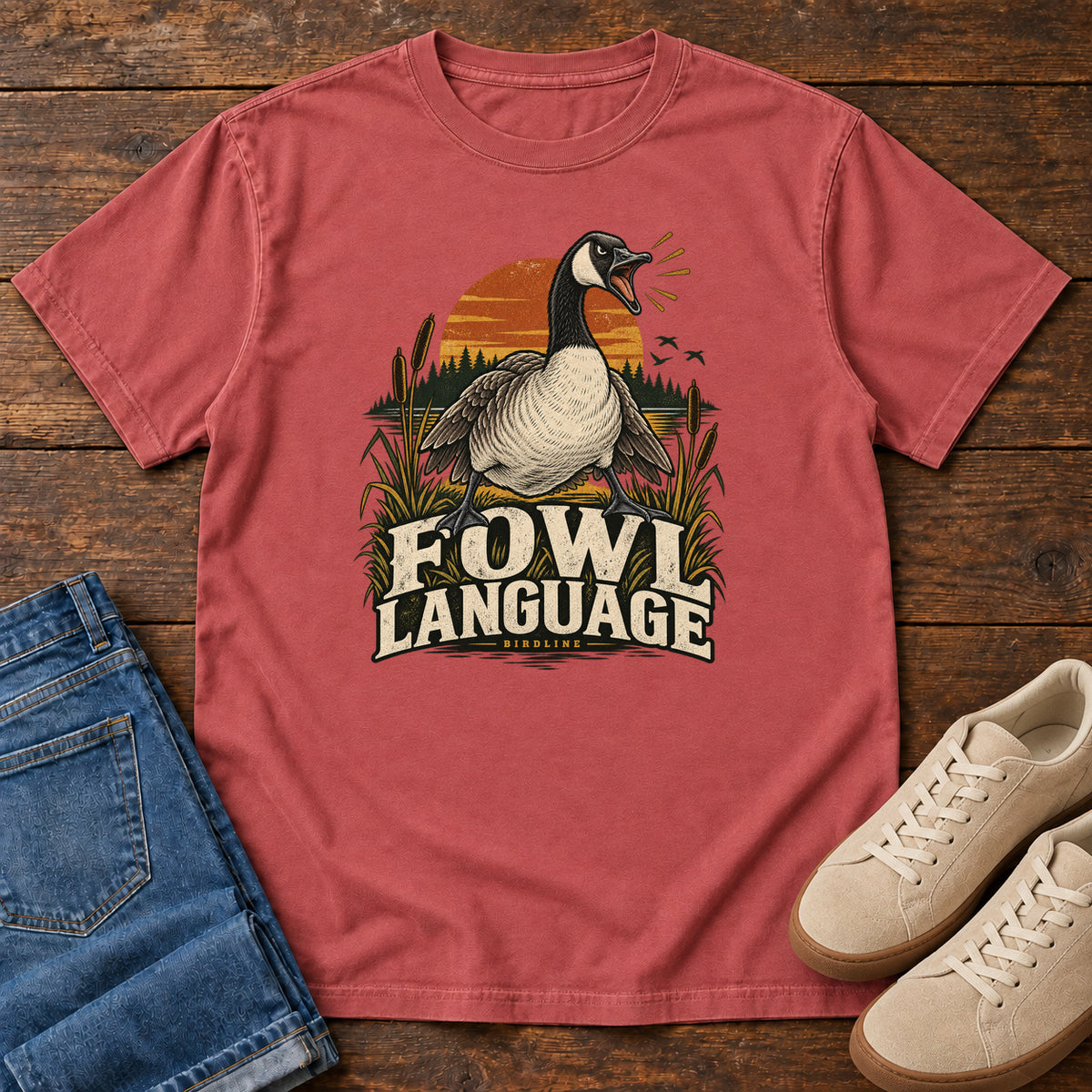 Crimson Fowl Language T-Shirt with an open-beaked goose among reeds against an orange sunset; front flat lay on wood.
