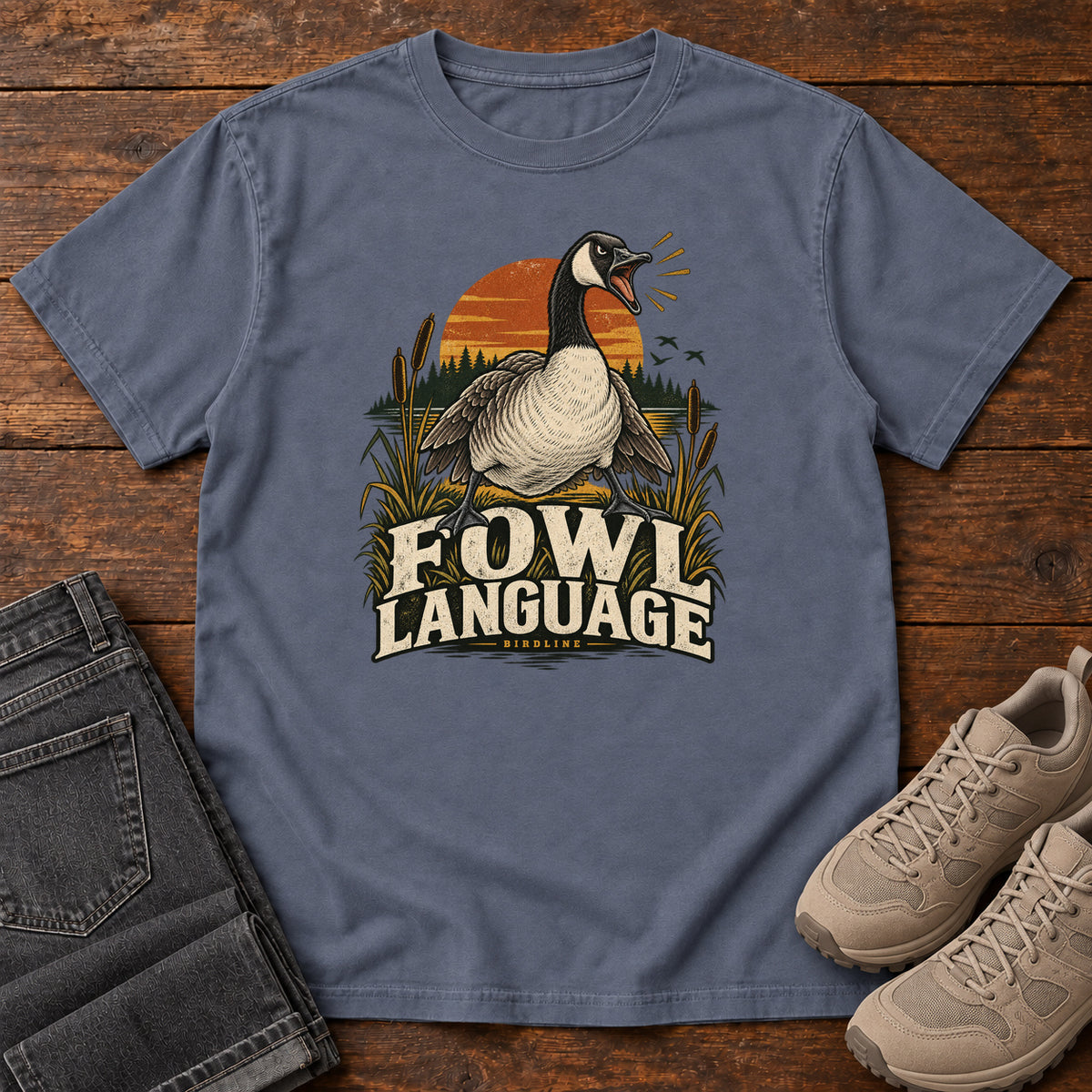 Denim Fowl Language T-Shirt with an open-beaked goose among reeds against an orange sunset; front flat lay on wood.