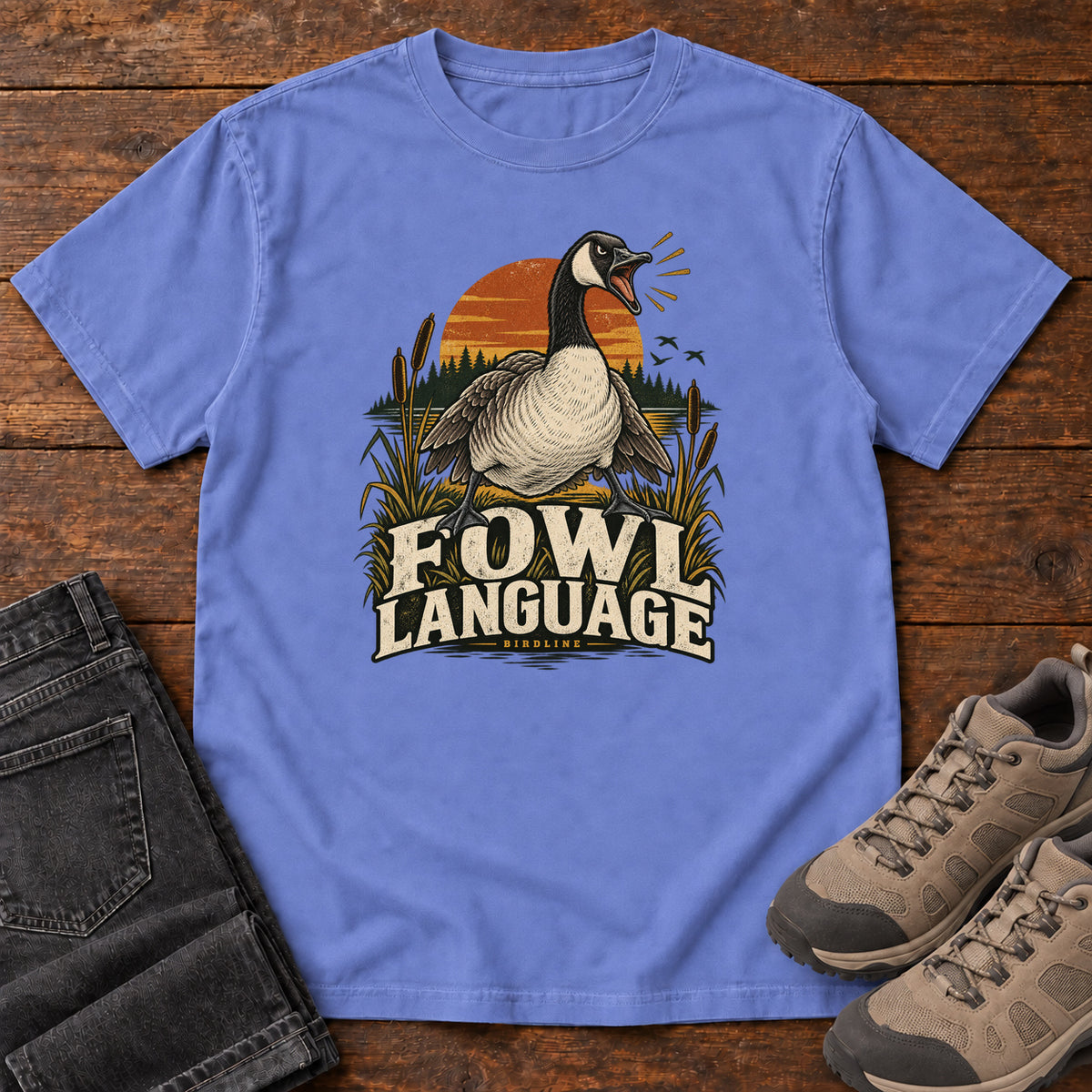Flo Blue Fowl Language T-Shirt with an open-beaked goose among reeds against an orange sunset; front flat lay on wood.