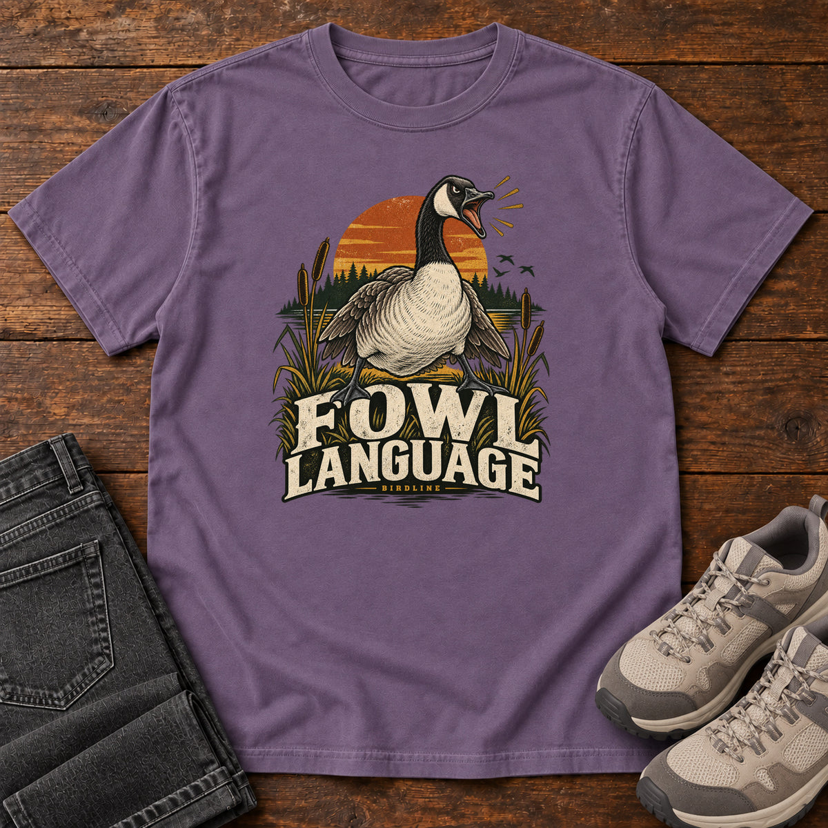 Grape Fowl Language T-Shirt with an open-beaked goose among reeds against an orange sunset; front flat lay on wood.