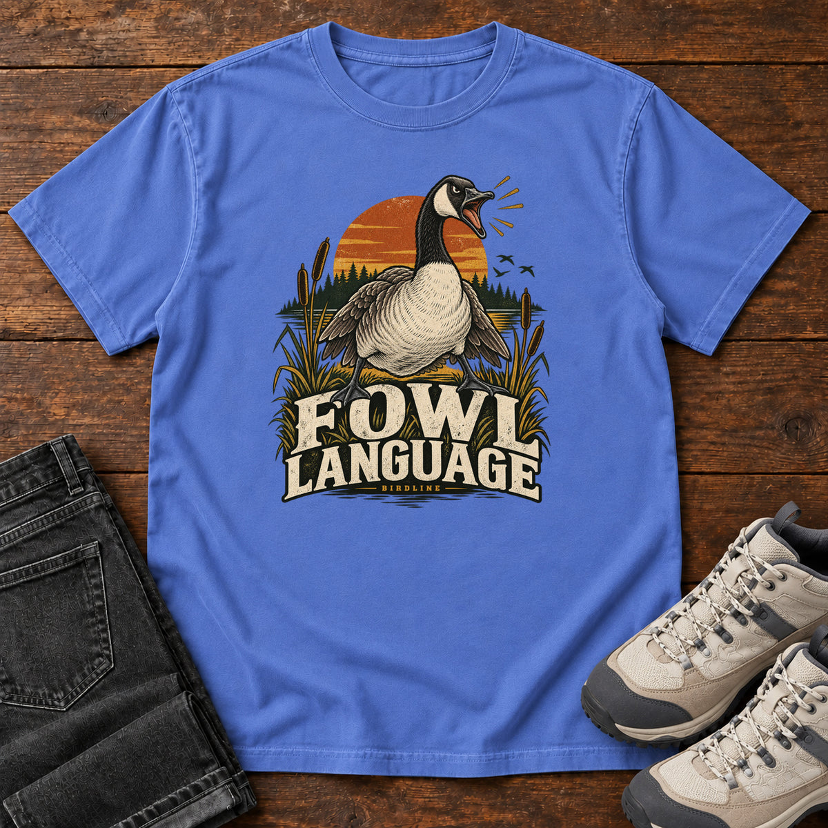 Mystic Blue Fowl Language T-Shirt with an open-beaked goose among reeds against an orange sunset; front flat lay on wood.