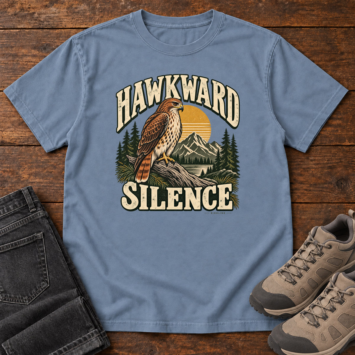 Blue Jean Hawkward Silence T-Shirt with a brown bird of prey perched above a mountain lake between cream Hawkward Silence lettering, front flat lay on wood.