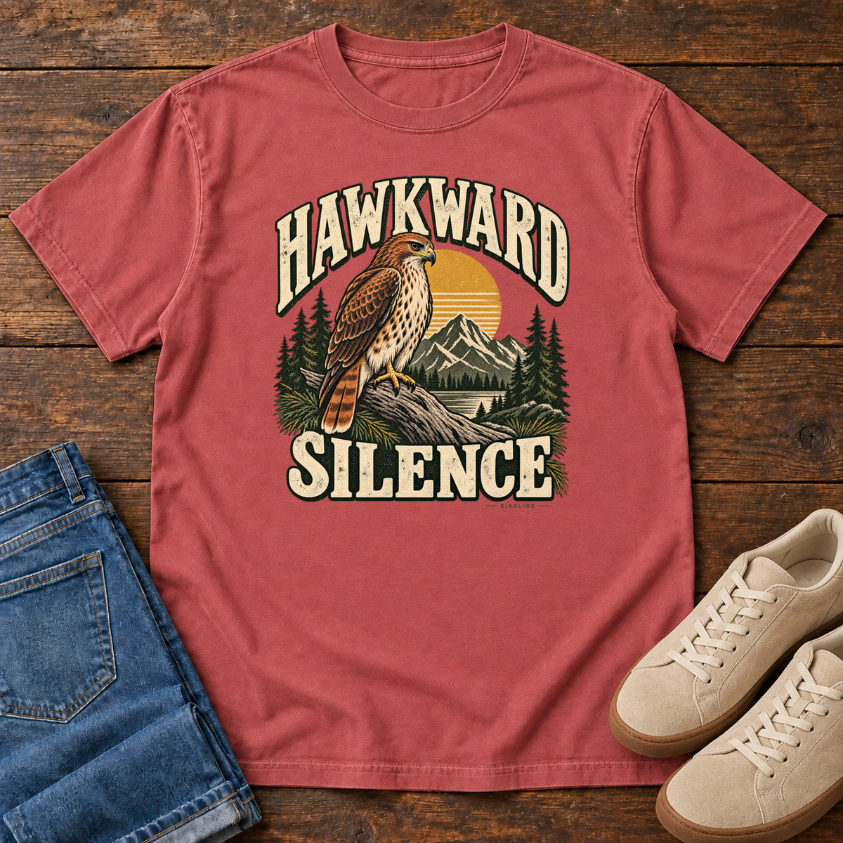 Crimson Hawkward Silence T-Shirt with a brown bird of prey perched above a mountain lake between cream Hawkward Silence lettering, front flat lay on wood.