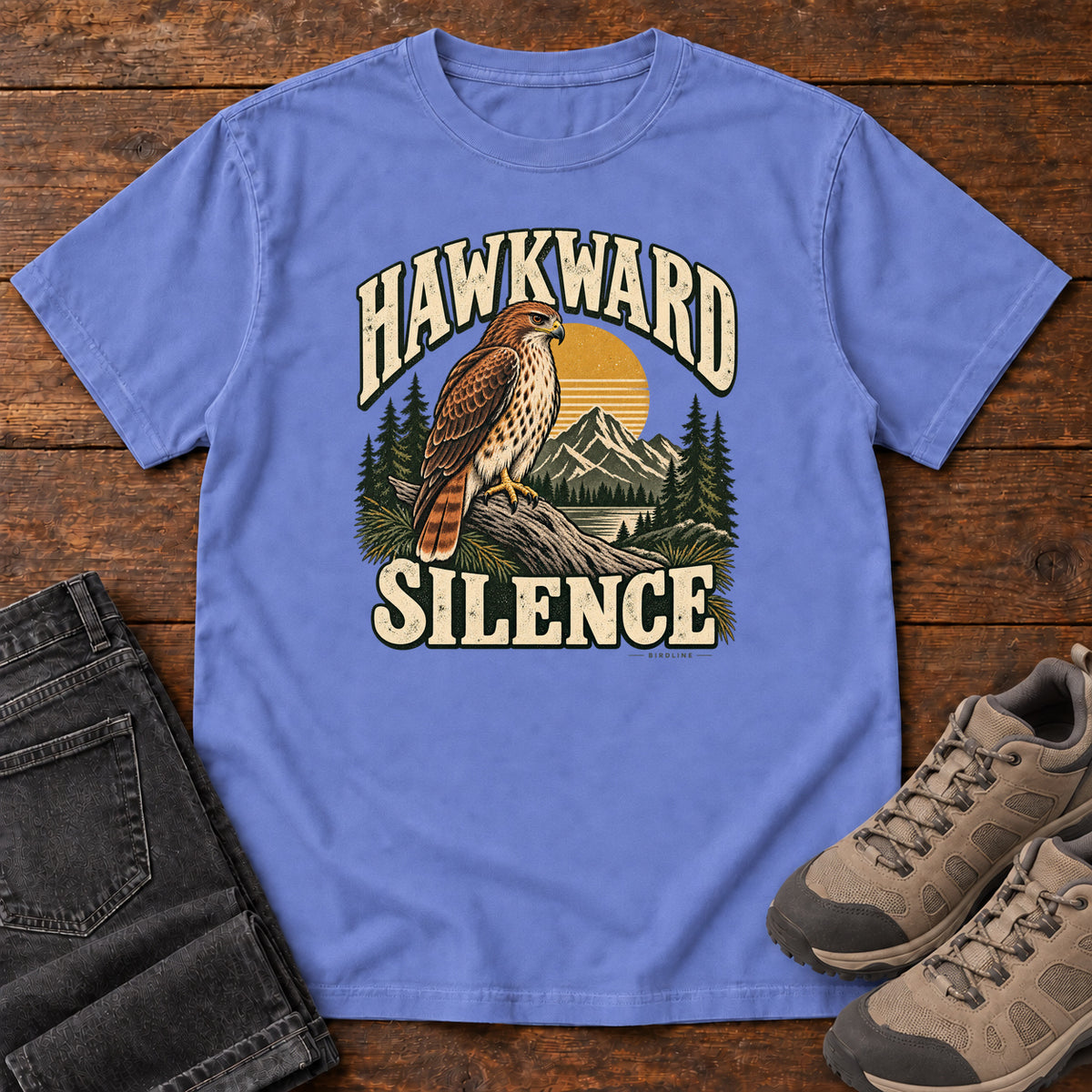 Flo Blue Hawkward Silence T-Shirt with a brown bird of prey perched above a mountain lake between cream Hawkward Silence lettering, front flat lay on wood.