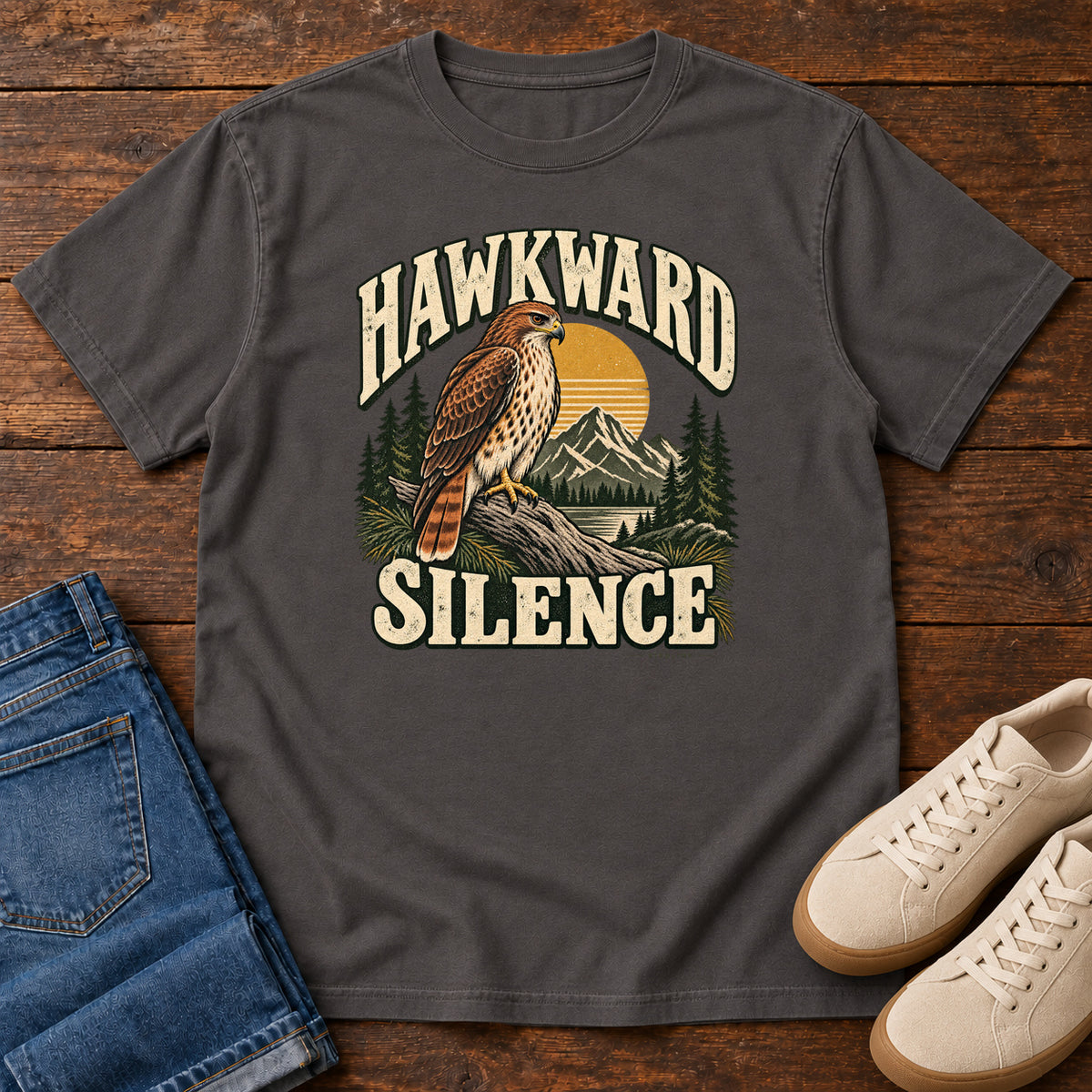 Graphite Hawkward Silence T-Shirt with a brown bird of prey perched above a mountain lake between cream Hawkward Silence lettering, front flat lay on wood.