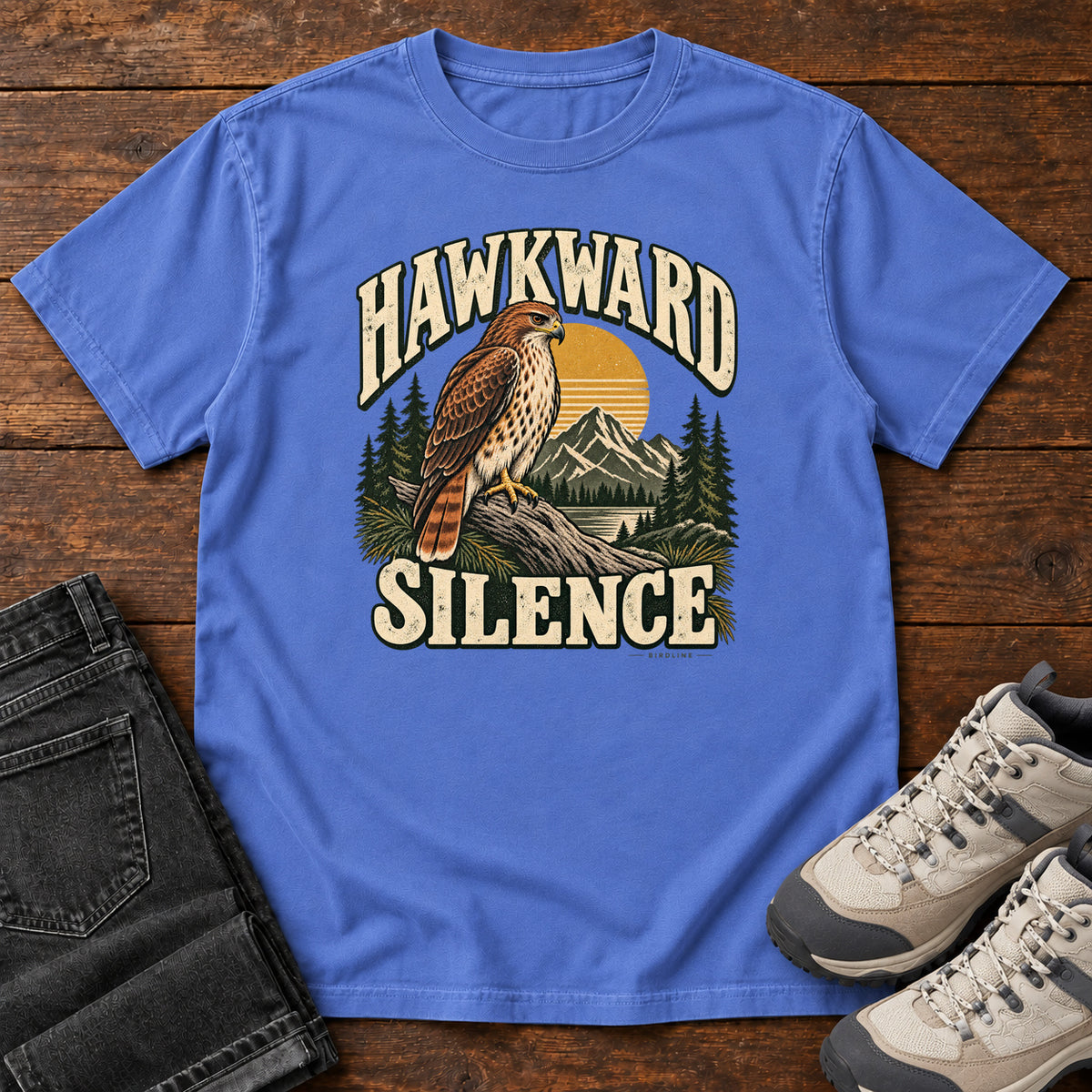Mystic Blue Hawkward Silence T-Shirt with a brown bird of prey perched above a mountain lake between cream Hawkward Silence lettering, front flat lay on wood.