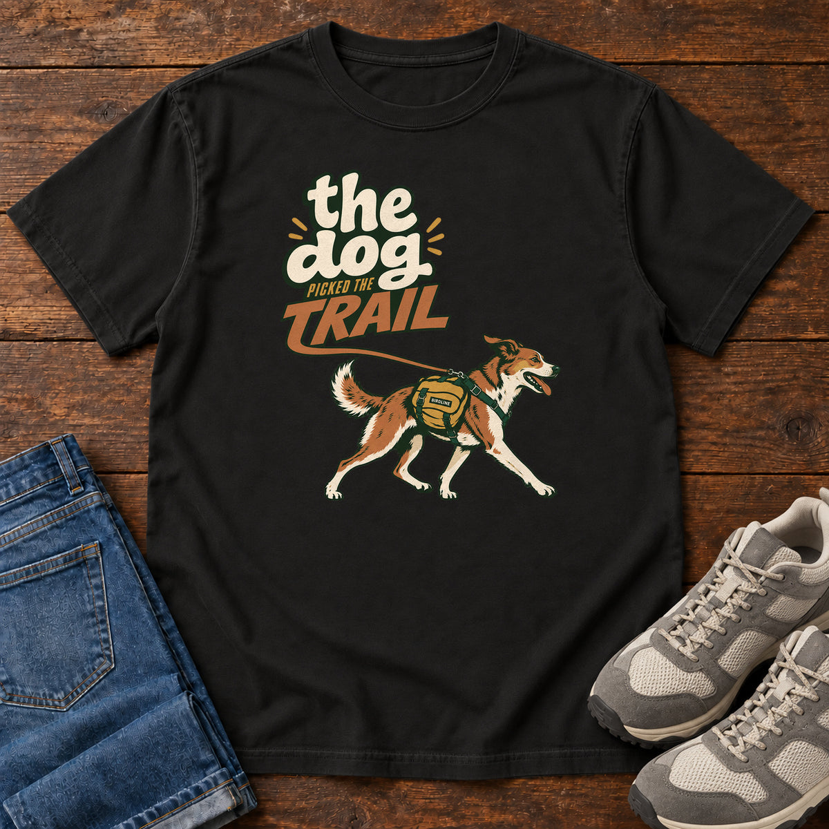 The Dog Picked the Trail T-Shirt in Black, front laid flat on wood, featuring a brown-and-white dog walking with a hiking pack beneath cream-and-orange lettering.