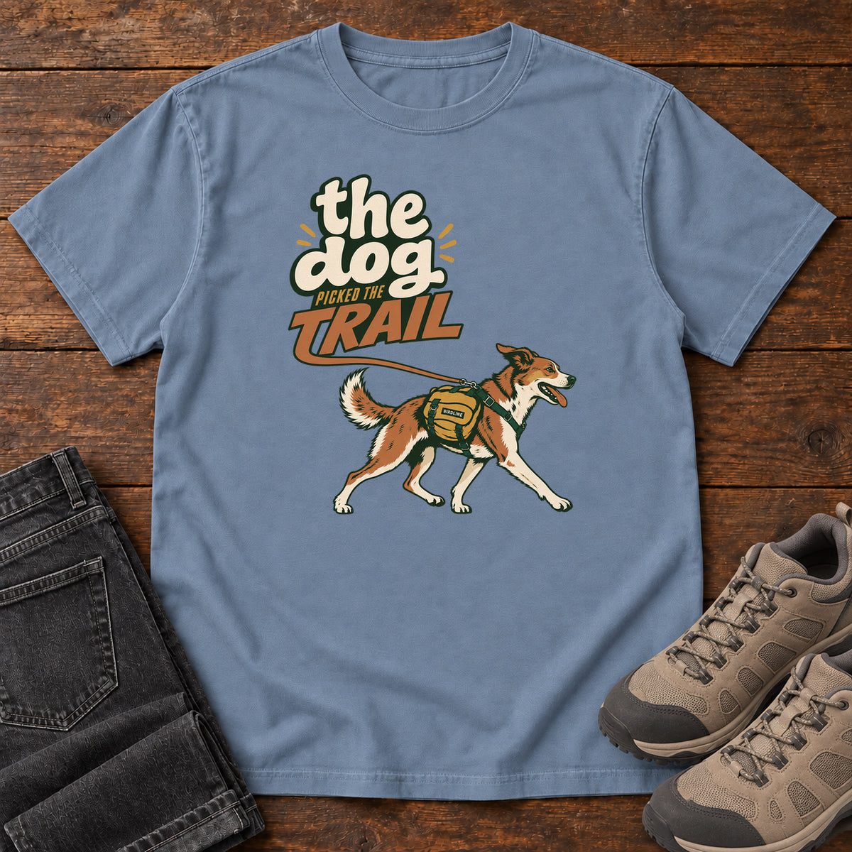 The Dog Picked the Trail T-Shirt in Blue Jean, front laid flat on wood, featuring a brown-and-white dog walking with a hiking pack beneath cream-and-orange lettering.