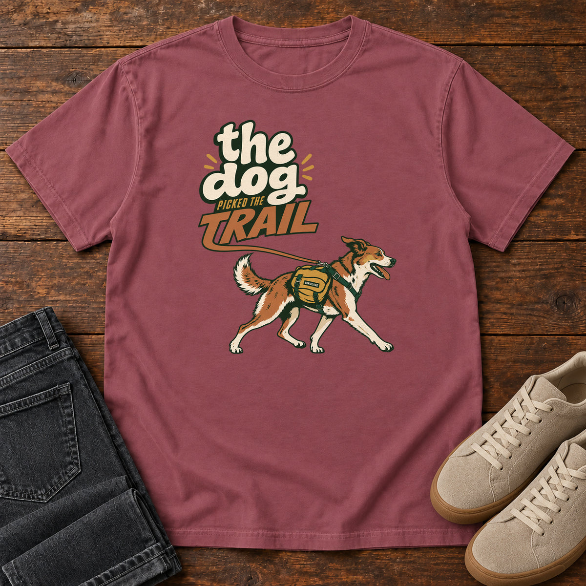 The Dog Picked the Trail T-Shirt in Brick, front laid flat on wood, featuring a brown-and-white dog walking with a hiking pack beneath cream-and-orange lettering.