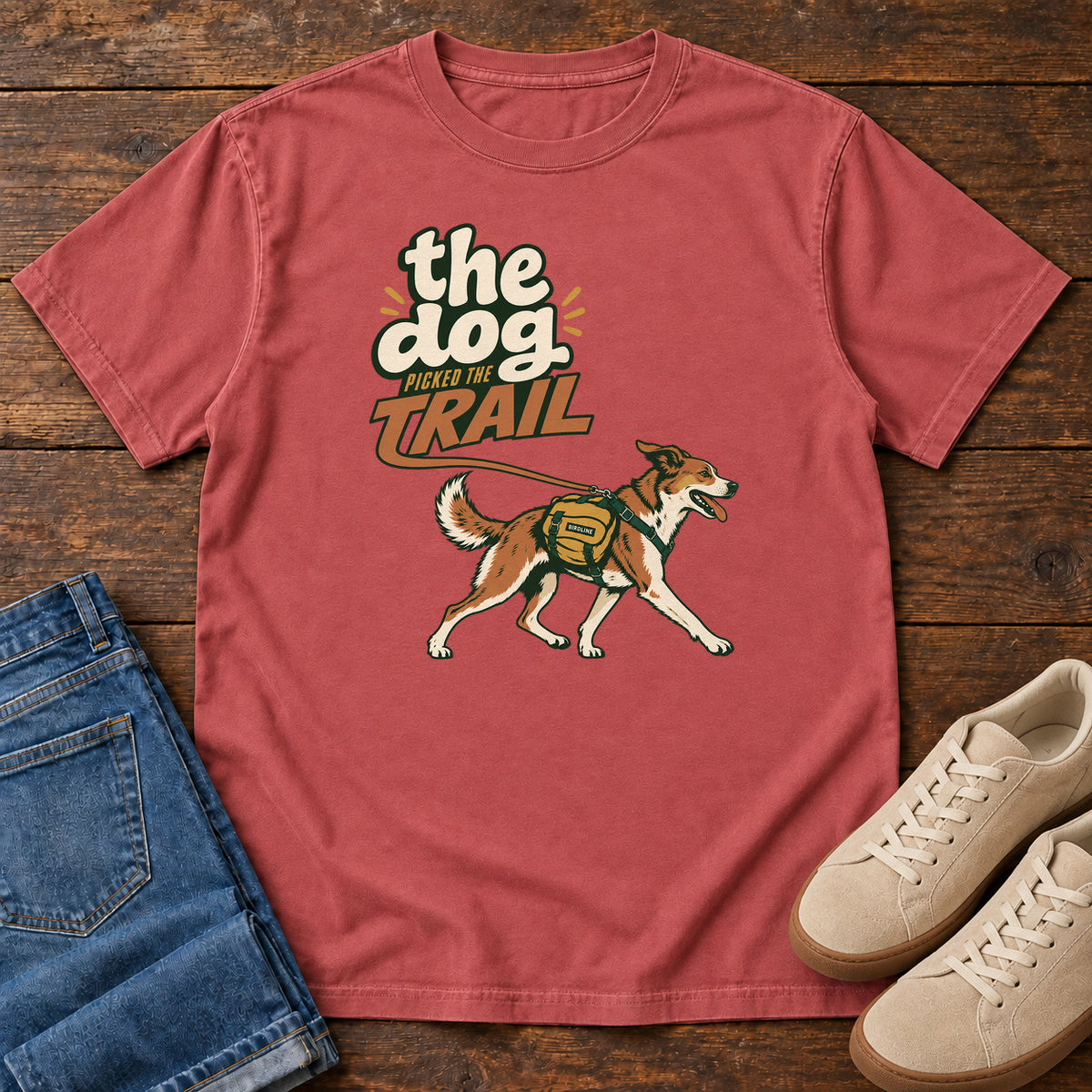 The Dog Picked the Trail T-Shirt in Crimson, front laid flat on wood, featuring a brown-and-white dog walking with a hiking pack beneath cream-and-orange lettering.