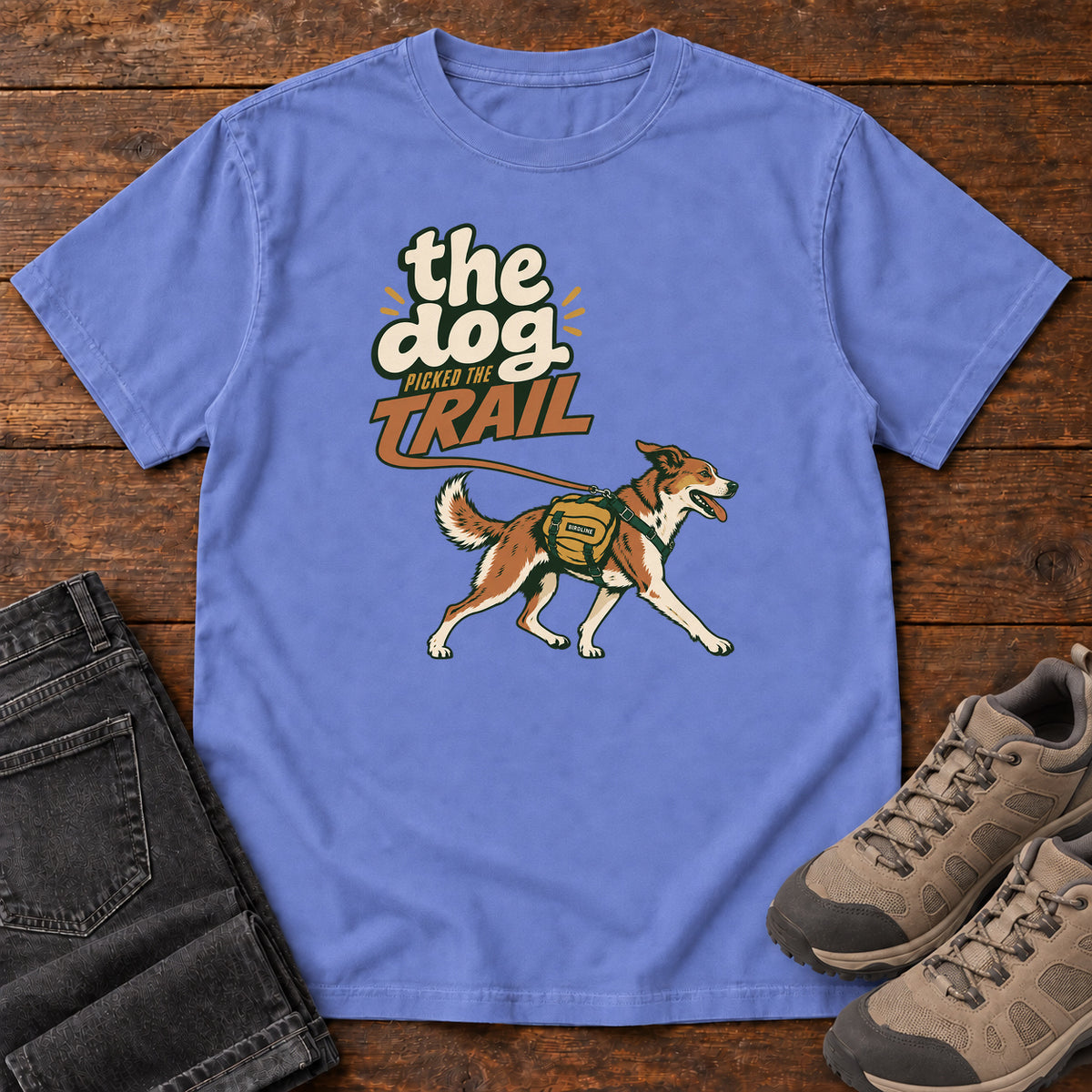 The Dog Picked the Trail T-Shirt in Flo Blue, front laid flat on wood, featuring a brown-and-white dog walking with a hiking pack beneath cream-and-orange lettering.