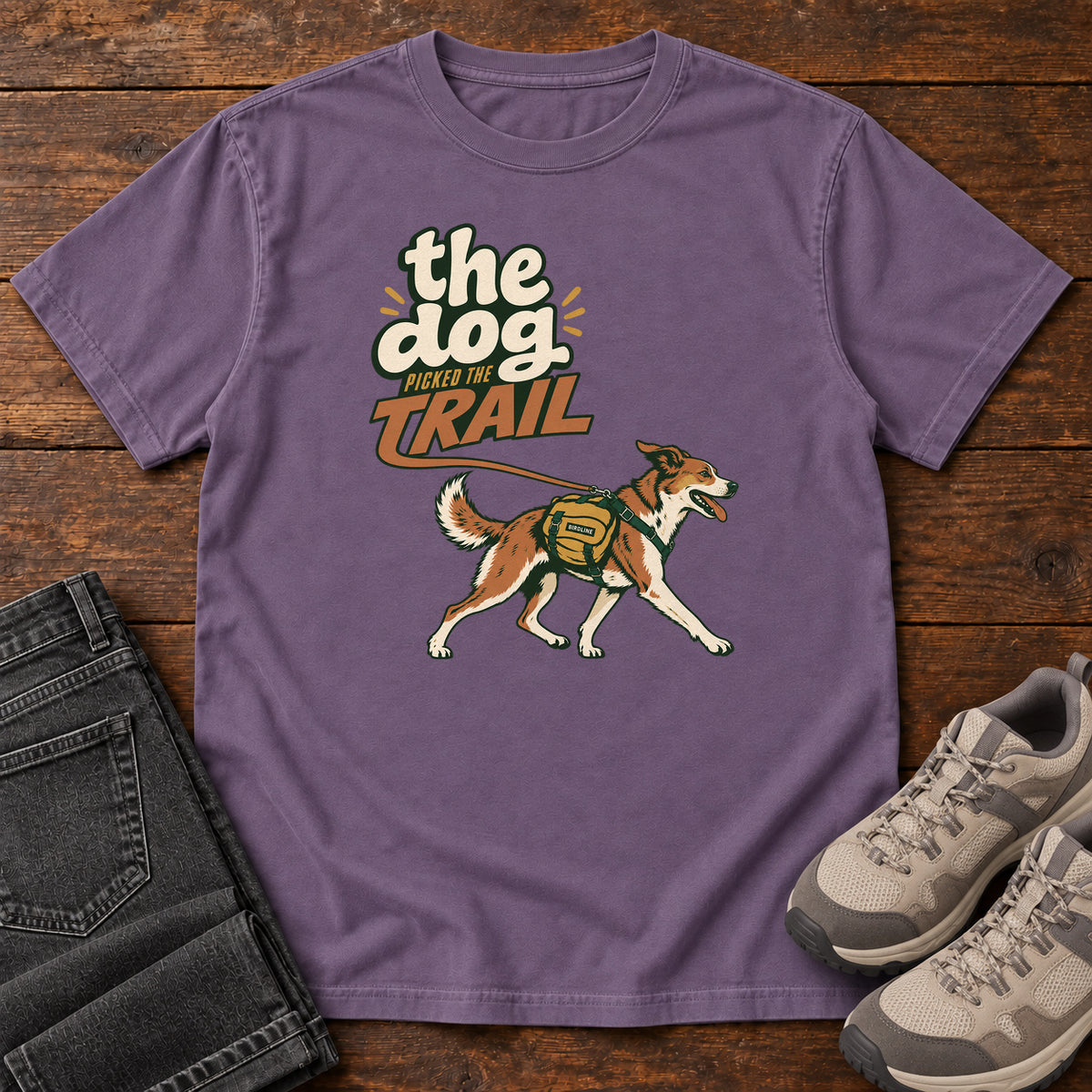 The Dog Picked the Trail T-Shirt in Grape, front laid flat on wood, featuring a brown-and-white dog walking with a hiking pack beneath cream-and-orange lettering.