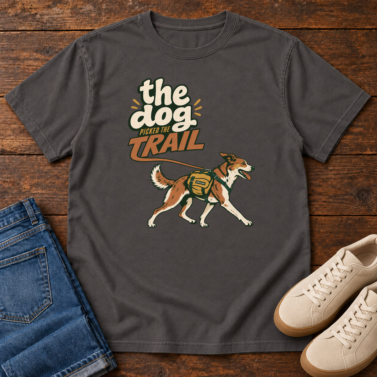 The Dog Picked the Trail T-Shirt in Graphite, front laid flat on wood, featuring a brown-and-white dog walking with a hiking pack beneath cream-and-orange lettering.