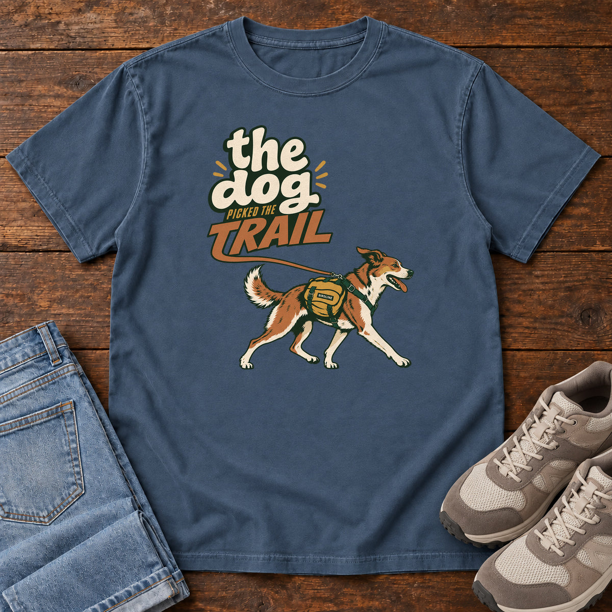The Dog Picked the Trail T-Shirt in Midnight, front laid flat on wood, featuring a brown-and-white dog walking with a hiking pack beneath cream-and-orange lettering.