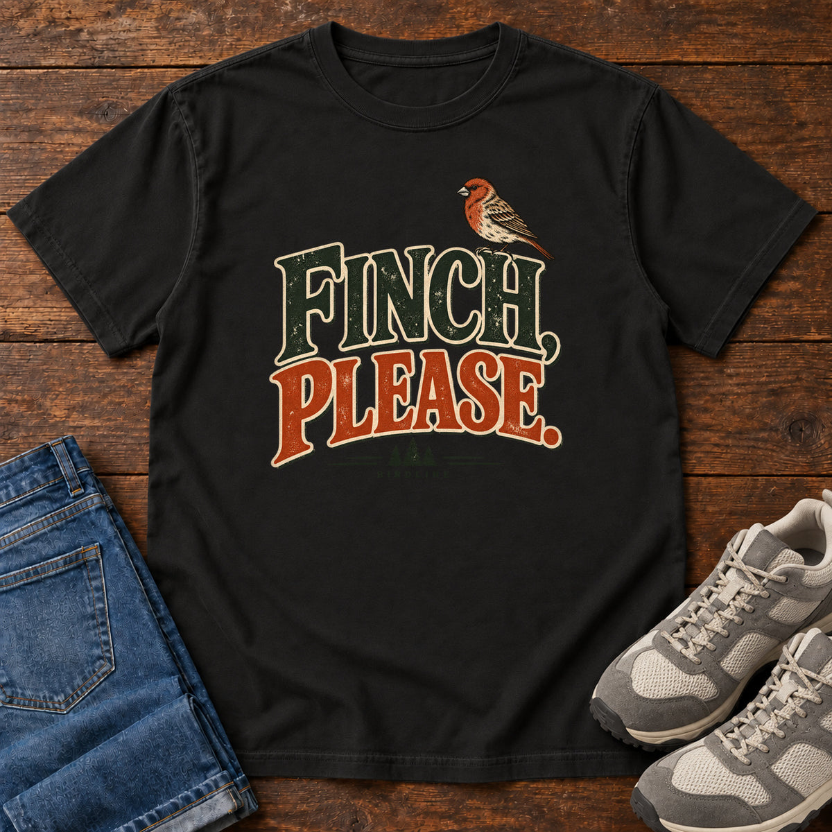 Black Finch, Please. T-Shirt with a small bird perched above cream-outlined green-and-orange lettering, front flat lay on wood.