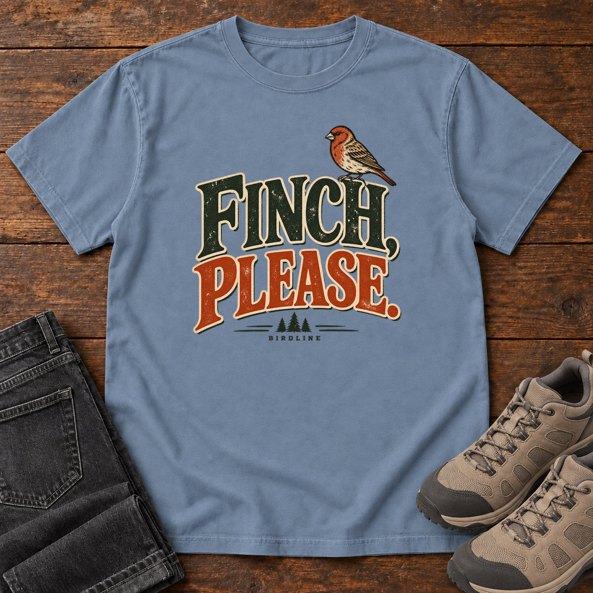 Blue Jean Finch, Please. T-Shirt with a small bird perched above cream-outlined green-and-orange lettering, front flat lay on wood.
