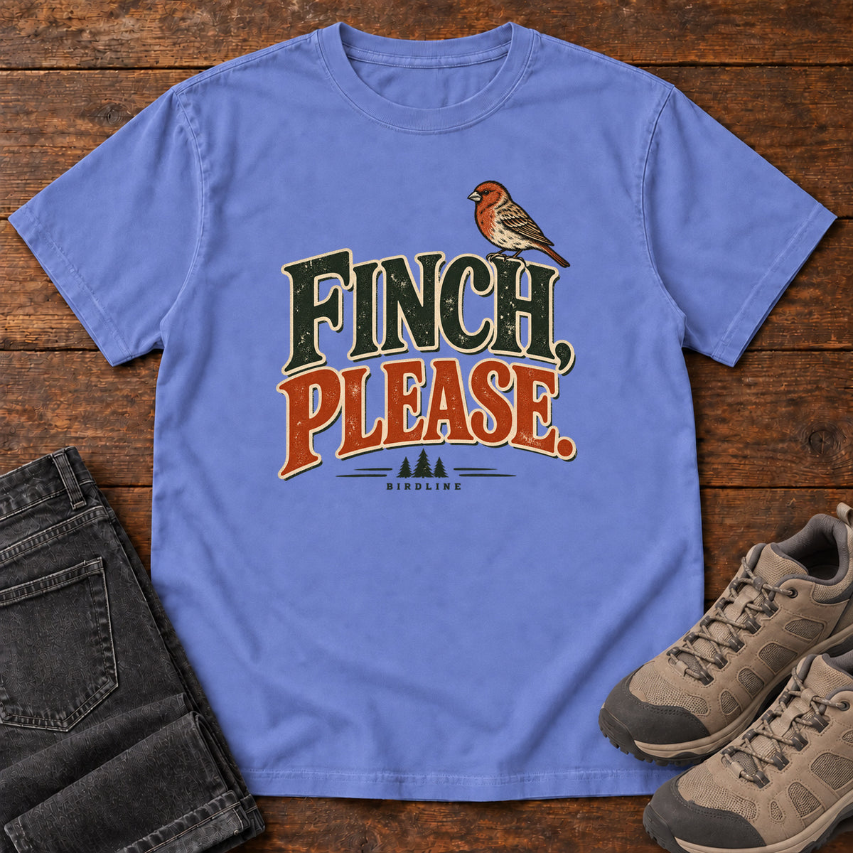 Flo Blue Finch, Please. T-Shirt with a small bird perched above cream-outlined green-and-orange lettering, front flat lay on wood.