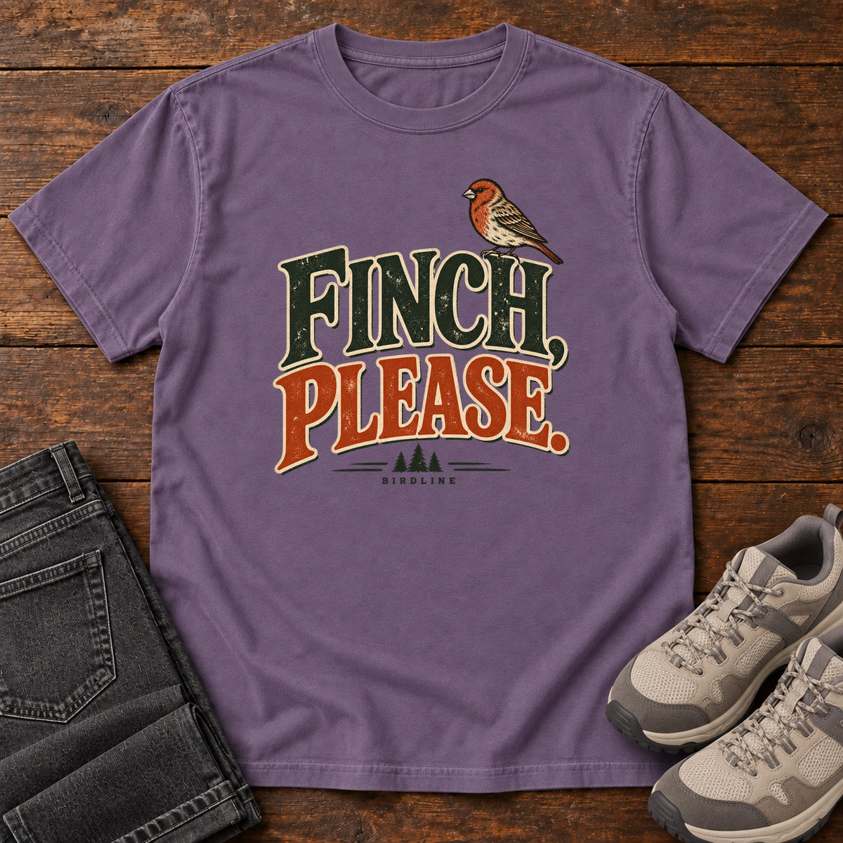 Grape Finch, Please. T-Shirt with a small bird perched above cream-outlined green-and-orange lettering, front flat lay on wood.