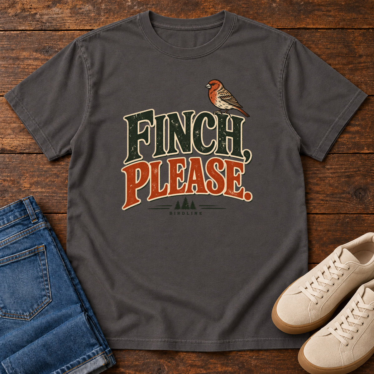 Graphite Finch, Please. T-Shirt with a small bird perched above cream-outlined green-and-orange lettering, front flat lay on wood.