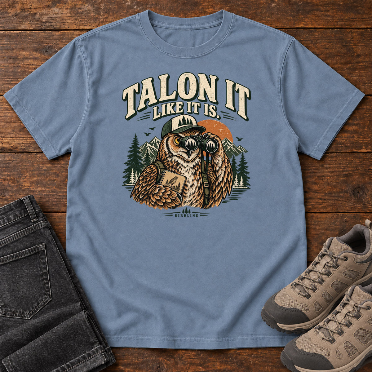 Blue Jean Talon It Like It Is. T-Shirt with an owl wearing a cap, looking through binoculars and holding a field notebook; front flat lay on wood.