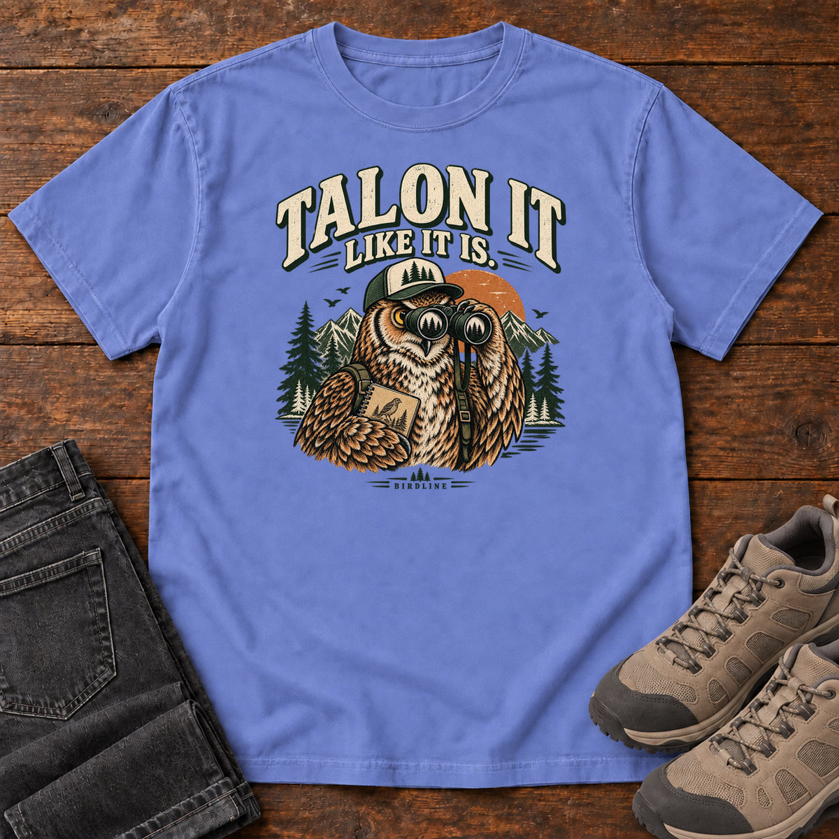 Flo Blue Talon It Like It Is. T-Shirt with an owl wearing a cap, looking through binoculars and holding a field notebook; front flat lay on wood.