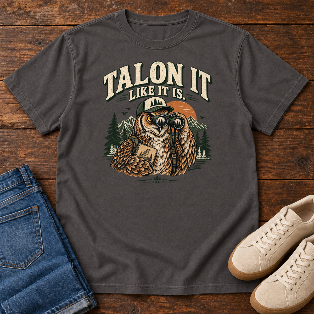 Graphite Talon It Like It Is. T-Shirt with an owl wearing a cap, looking through binoculars and holding a field notebook; front flat lay on wood.