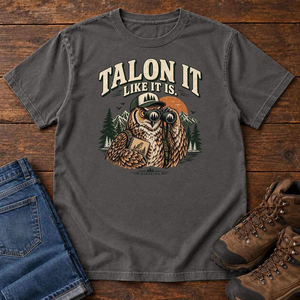Pepper Talon It Like It Is. T-Shirt with an owl wearing a cap, looking through binoculars and holding a field notebook; front flat lay on wood.