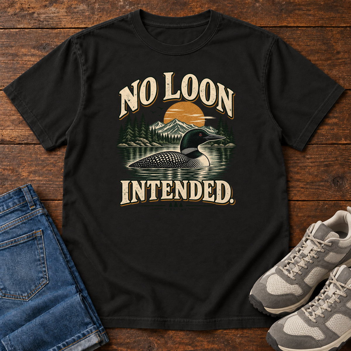 Black No Loon Intended T-Shirt with a black-and-white waterbird on a mountain lake beneath an orange sun and cream lettering, front flat lay on wood.