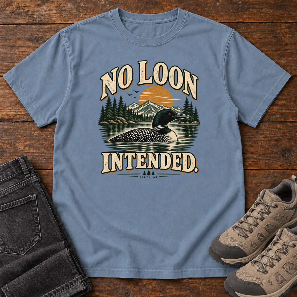 Blue Jean No Loon Intended T-Shirt with a black-and-white waterbird on a mountain lake beneath an orange sun and cream lettering, front flat lay on wood.