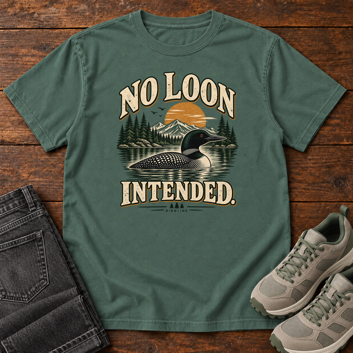 Blue Spruce No Loon Intended T-Shirt with a black-and-white waterbird on a mountain lake beneath an orange sun and cream lettering, front flat lay on wood.