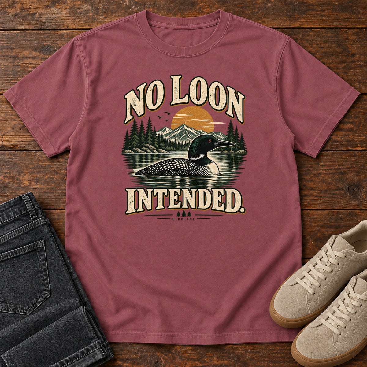 Brick No Loon Intended T-Shirt with a black-and-white waterbird on a mountain lake beneath an orange sun and cream lettering, front flat lay on wood.