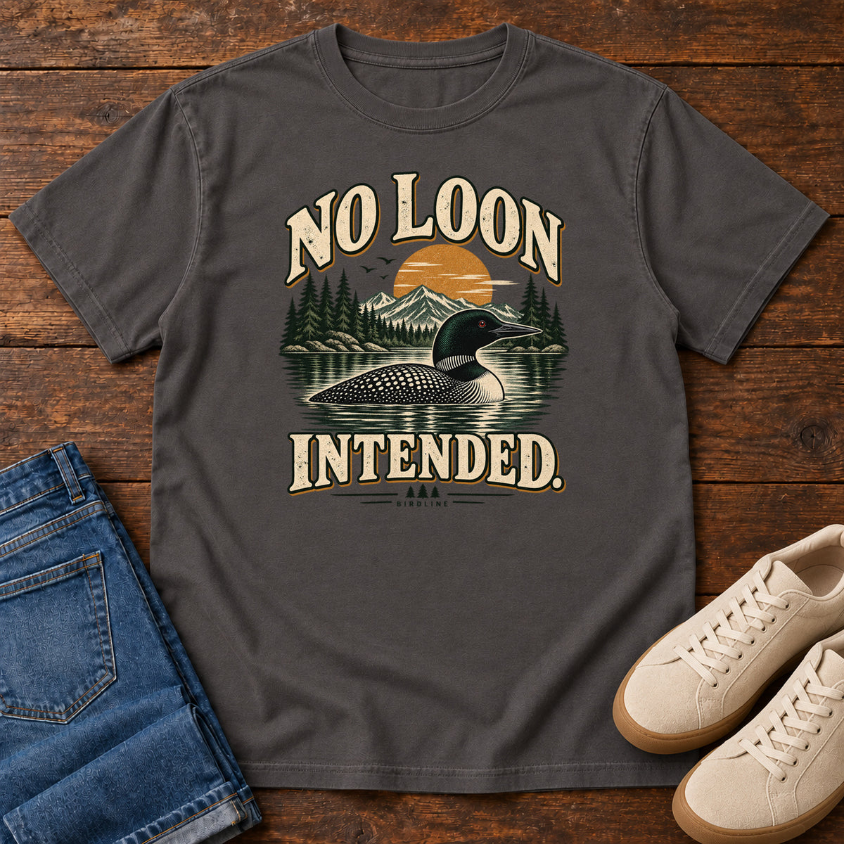 Graphite No Loon Intended T-Shirt with a black-and-white waterbird on a mountain lake beneath an orange sun and cream lettering, front flat lay on wood.