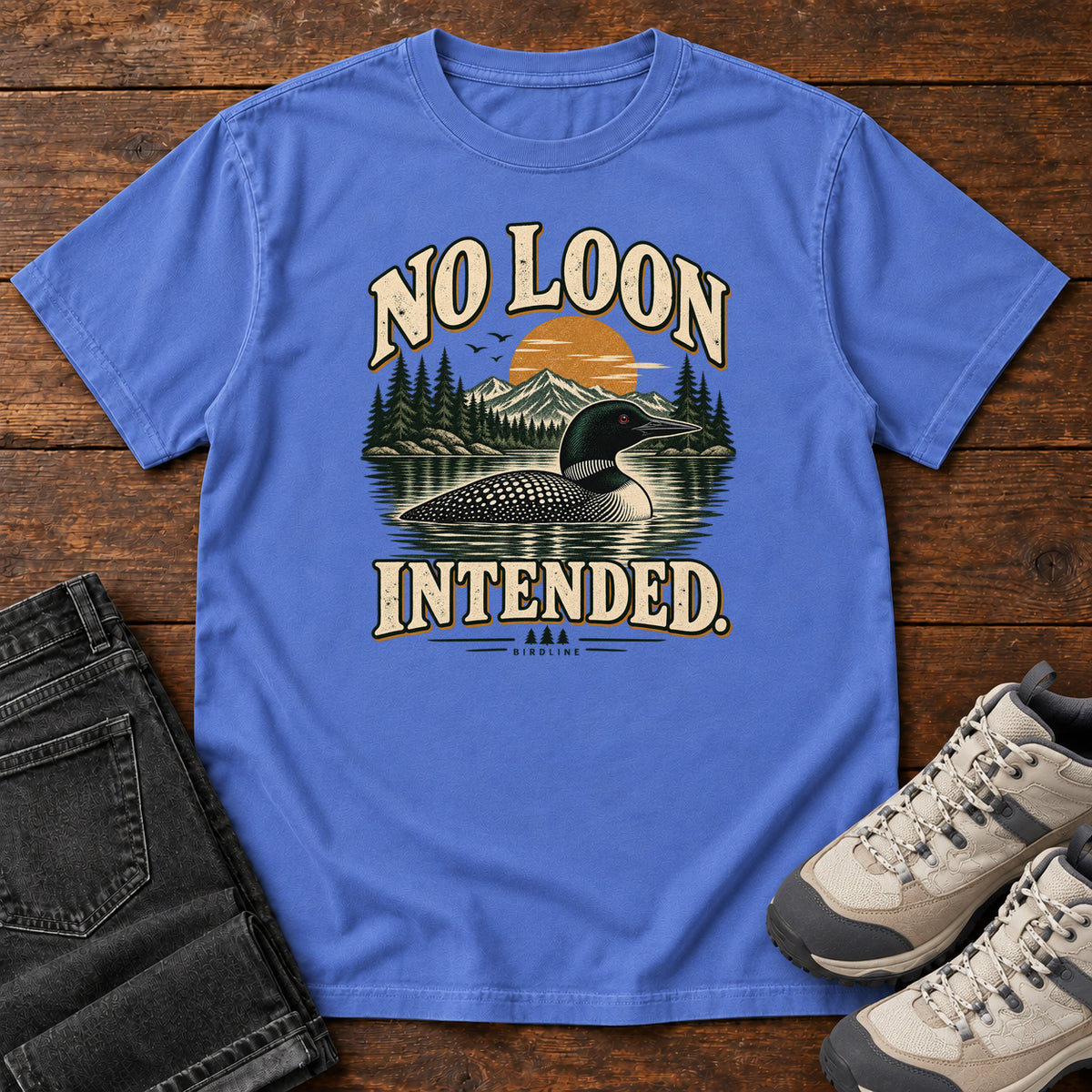 Mystic Blue No Loon Intended T-Shirt with a black-and-white waterbird on a mountain lake beneath an orange sun and cream lettering, front flat lay on wood.