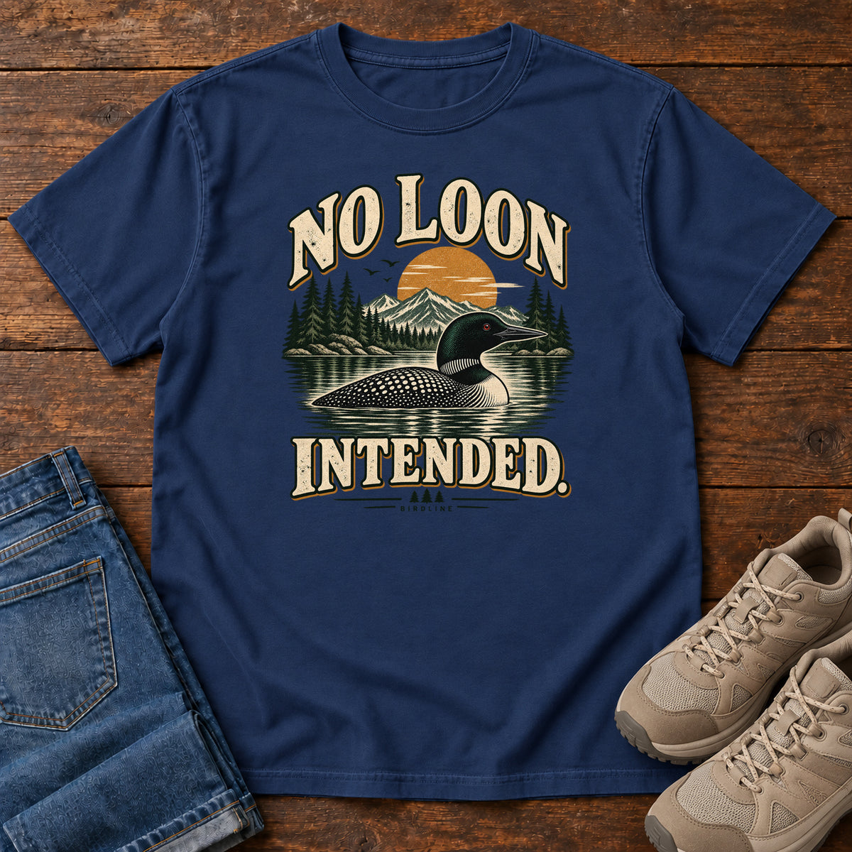 True Navy No Loon Intended T-Shirt with a black-and-white waterbird on a mountain lake beneath an orange sun and cream lettering, front flat lay on wood.