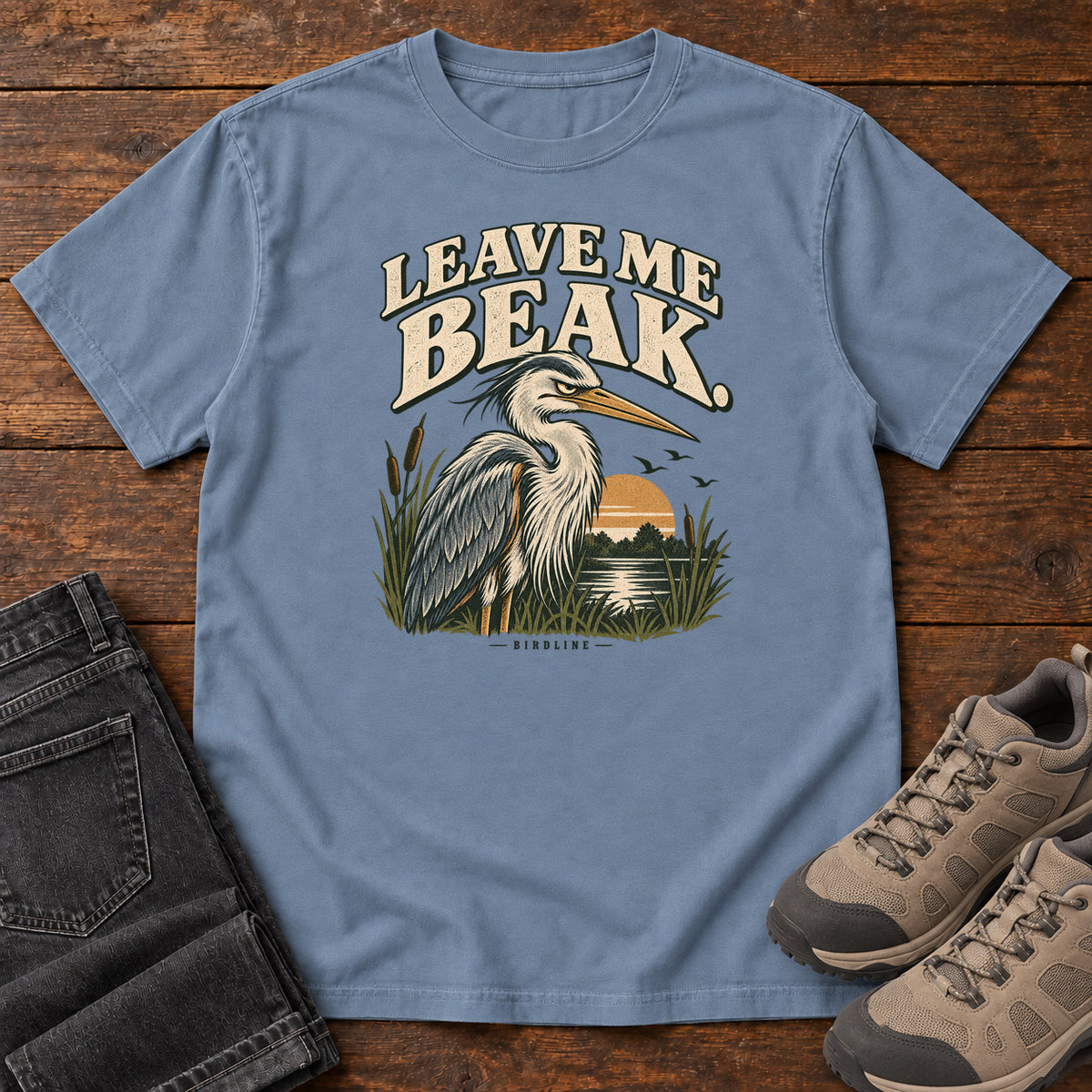 Leave Me Beak T-Shirt in Blue Jean, front laid flat on wood, featuring a gray, long-billed wading bird among reeds beside a sunset, beneath cream lettering.