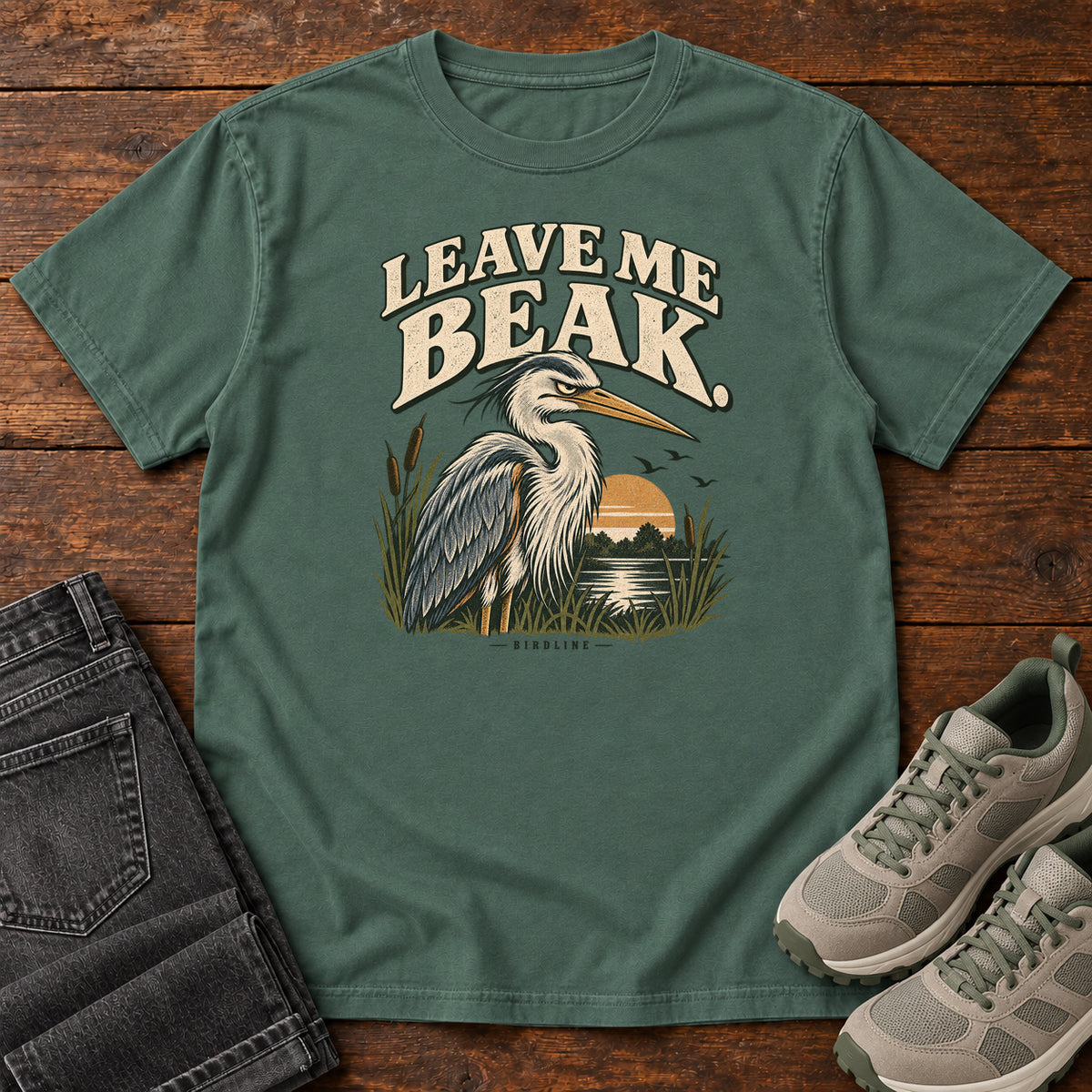 Leave Me Beak T-Shirt in Blue Spruce, front laid flat on wood, featuring a gray, long-billed wading bird among reeds beside a sunset, beneath cream lettering.