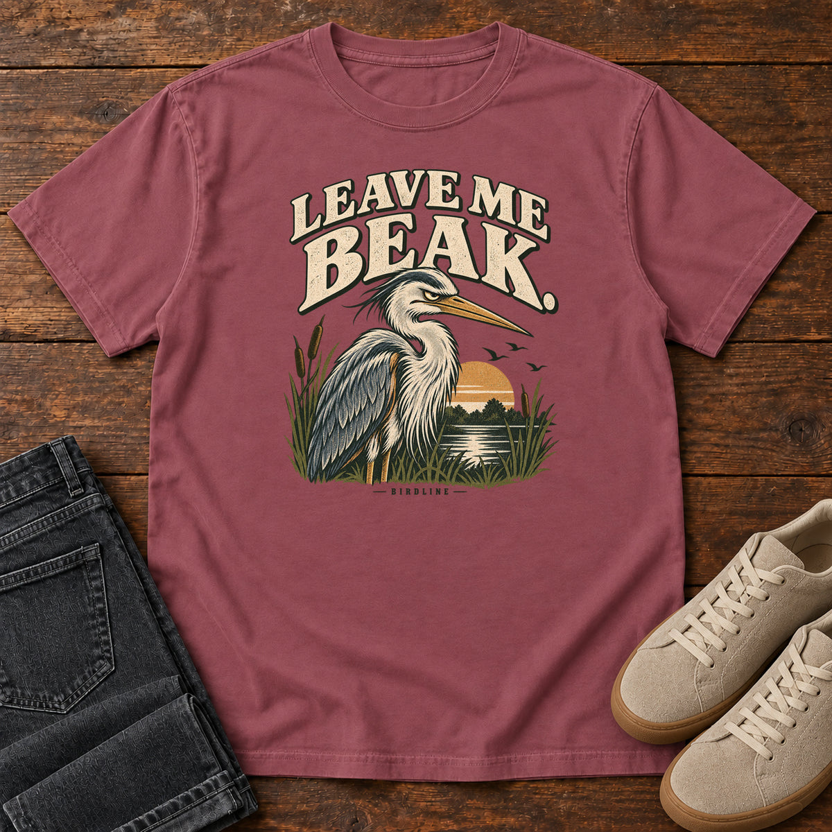 Leave Me Beak T-Shirt in Brick, front laid flat on wood, featuring a gray, long-billed wading bird among reeds beside a sunset, beneath cream lettering.